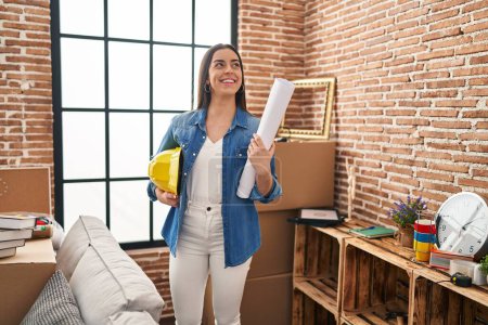 Photo for Young beautiful hispanic woman architect smiling confident holding hardhat and blueprints at new home - Royalty Free Image
