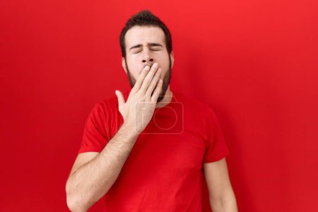 Téléchargez les photos : Jeune homme hispanique portant un t-shirt rouge occasionnel ennuyé bâillant fatigué couvrant la bouche avec la main. agitation et somnolence. - en image libre de droit