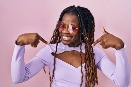 Foto de Mujer africana con el pelo trenzado de pie sobre fondo rosa mirando confiado con sonrisa en la cara, señalándose con los dedos orgullosos y felices. - Imagen libre de derechos