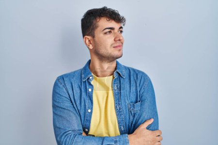 Téléchargez les photos : Jeune homme hispanique debout sur fond bleu regardant du côté avec les bras croisés convaincu et confiant - en image libre de droit
