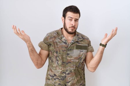 Téléchargez les photos : Jeune homme hispanique portant uniforme de camouflage de l'armée expression désemparée et confuse avec les bras et les mains levées. concept de doute. - en image libre de droit