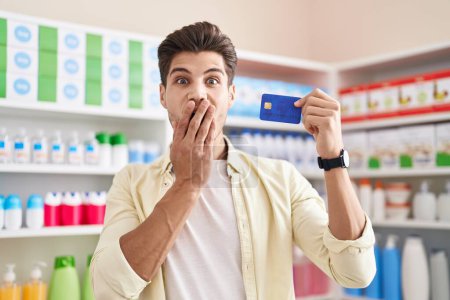 Téléchargez les photos : Jeune homme hispanique à la pharmacie pharmacie tenant carte de crédit couvrant la bouche avec la main, choqué et effrayé par l'erreur. expression surprise - en image libre de droit