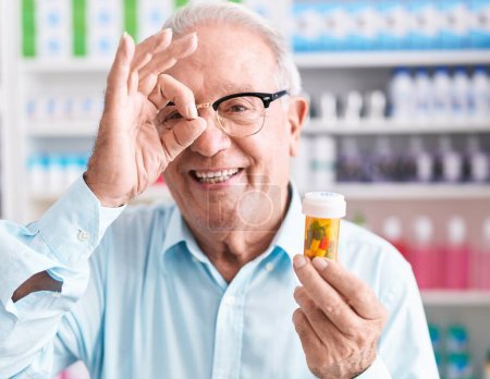 Téléchargez les photos : Homme âgé avec les cheveux gris tenant pilules à la pharmacie souriant heureux faire ok signe avec la main sur les yeux en regardant à travers les doigts - en image libre de droit