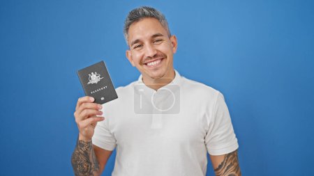 Téléchargez les photos : Jeune homme hispanique souriant confiant tenant passeport sur fond bleu isolé - en image libre de droit