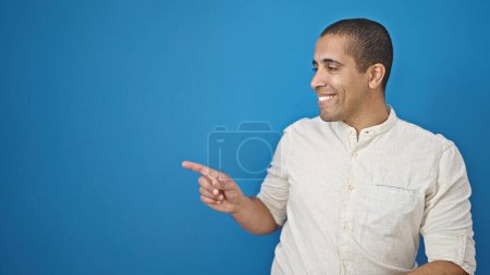 Foto de Joven hombre hispano sonriendo señalando a un lado sobre un fondo azul aislado - Imagen libre de derechos