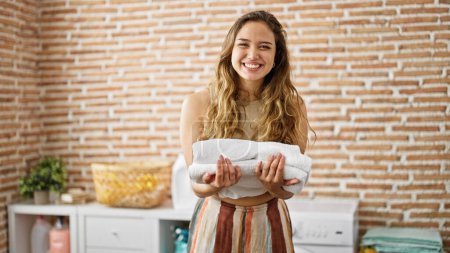 Foto de Joven hermosa mujer hispana sonriendo confiada sosteniendo toallas dobladas en la lavandería - Imagen libre de derechos