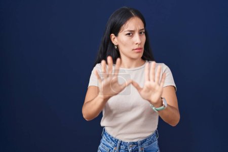Foto de Mujer hispana joven de pie sobre fondo azul alejando las palmas de las manos mostrando rechazo y negación con miedo y expresión repugnante. parada y prohibido. - Imagen libre de derechos