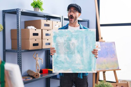 Foto de Hombre hispano con barba sosteniendo el lienzo en el estudio en la cara de choque, mirando escéptico y sarcástico, sorprendido con la boca abierta - Imagen libre de derechos