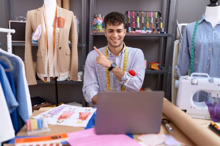 Téléchargez les photos : Jeune homme hispanique couturière designer à l'aide d'un ordinateur portable souriant pointant joyeusement avec la main et le doigt sur le côté - en image libre de droit