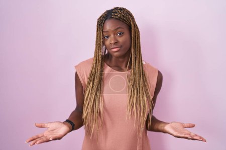 Téléchargez les photos : Femme afro-américaine avec des cheveux tressés debout sur fond rose désemparé et confondu avec les bras ouverts, aucune idée concept. - en image libre de droit
