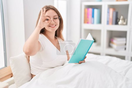 Foto de Joven mujer caucásica sentada en la cama en casa leyendo un libro sonriendo feliz haciendo ok signo con la mano en el ojo mirando a través de los dedos - Imagen libre de derechos