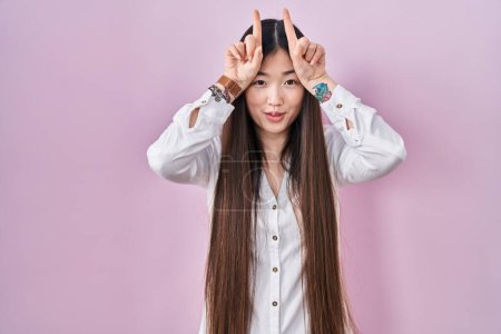 Photo for Chinese young woman standing over pink background doing funny gesture with finger over head as bull horns - Royalty Free Image