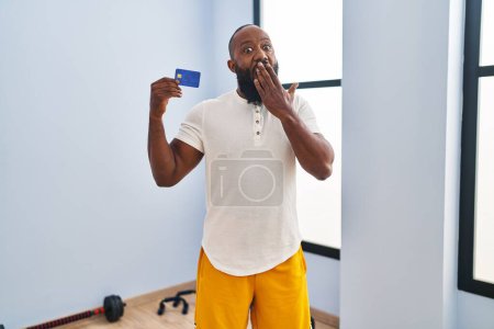Foto de Hombre afroamericano usando ropa deportiva con tarjeta de crédito cubriendo la boca con la mano, sorprendido y temeroso de equivocarse. expresión sorprendida - Imagen libre de derechos