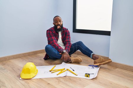 Photo for African american man sitting on the floor at new home looking at blueprints sticking tongue out happy with funny expression. emotion concept. - Royalty Free Image