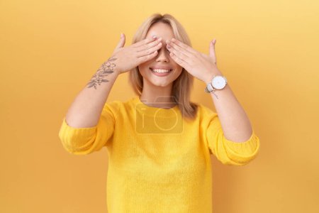 Téléchargez les photos : Jeune femme caucasienne portant un pull jaune couvrant les yeux avec les mains souriantes gaies et drôles. concept aveugle. - en image libre de droit