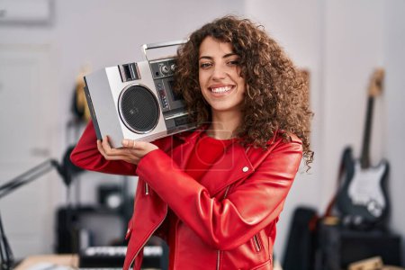 Téléchargez les photos : Femme hispanique aux cheveux bouclés tenant boombox, écoutant de la musique à l'air positif et heureux debout et souriant avec un sourire confiant montrant les dents - en image libre de droit