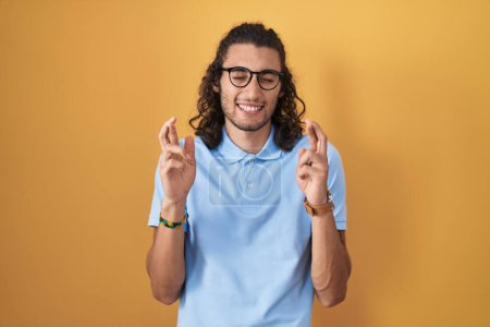 Téléchargez les photos : Jeune homme hispanique debout sur fond jaune geste doigt croisé souriant avec espoir et les yeux fermés. chance et concept superstitieux. - en image libre de droit