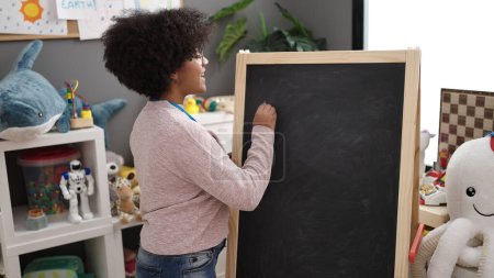 Photo for Young african american woman preschool teacher smiling confident writing on blackboard at kindergarten - Royalty Free Image