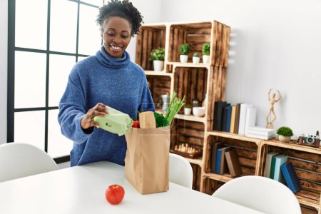 Téléchargez les photos : Femme afro-américaine souriante confiante tenant de la nourriture de sac d'épicerie à la maison - en image libre de droit