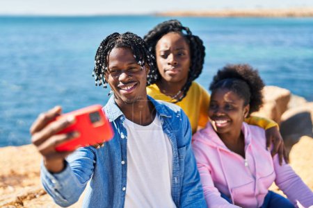 Téléchargez les photos : Des amis afro-américains faisant du selfie par le smartphone assis sur le rocher au bord de la mer - en image libre de droit