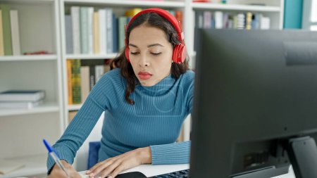 Téléchargez les photos : Jeune belle étudiante hispanique utilisant l'ordinateur prenant des notes portant des écouteurs à l'université salle de classe - en image libre de droit