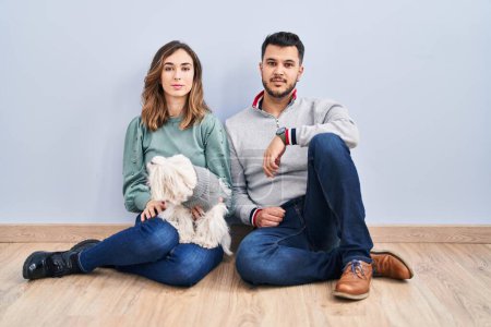 Téléchargez les photos : Jeune couple hispanique assis sur le sol avec chien détendu avec une expression sérieuse sur le visage. simple et naturel regardant la caméra. - en image libre de droit
