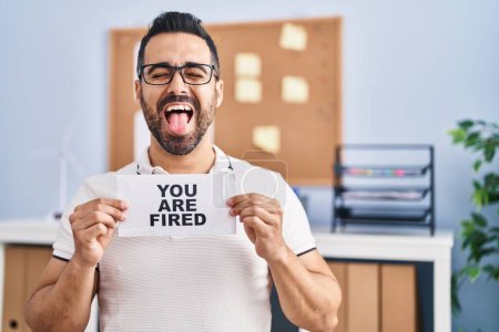 Photo for Young hispanic man with beard holding you are fired banner at the office sticking tongue out happy with funny expression. - Royalty Free Image