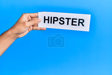 Foto de Mano de hombre caucásico sosteniendo papel con palabra hipster sobre fondo azul aislado - Imagen libre de derechos