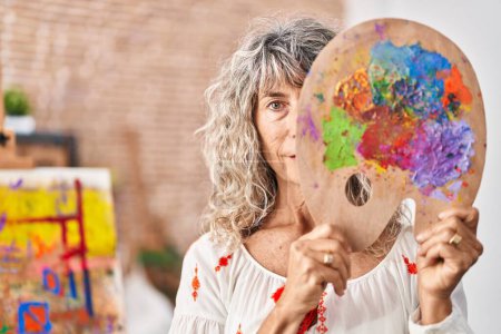 Photo for Middle age woman holding painter palette covering face relaxed with serious expression on face. simple and natural looking at the camera. - Royalty Free Image
