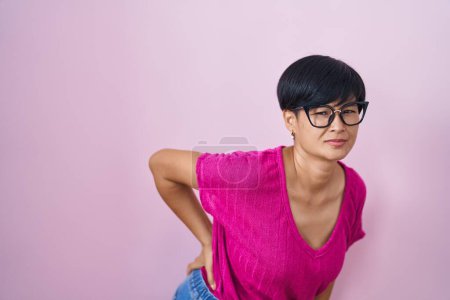 Téléchargez les photos : Jeune femme asiatique aux cheveux courts debout sur fond rose souffrant de maux de dos, toucher le dos avec la main, douleur musculaire - en image libre de droit