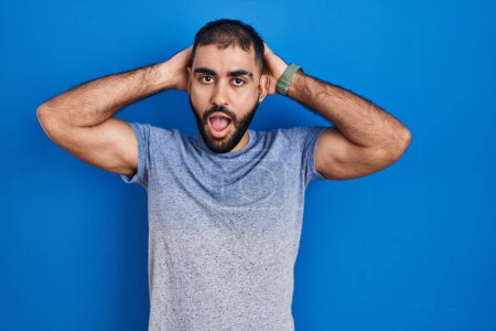 Téléchargez les photos : Moyen-Orient homme avec barbe debout sur fond bleu fou et effrayé avec les mains sur la tête, peur et surpris de choc avec la bouche ouverte - en image libre de droit
