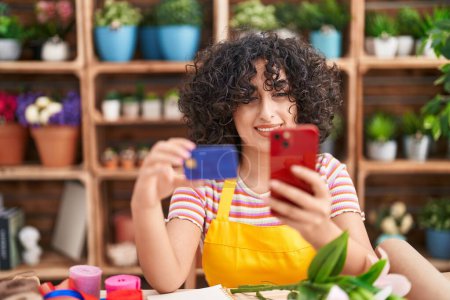 Téléchargez les photos : Jeune fleuriste moyen-orientale utilisant un smartphone tenant une carte de crédit chez le fleuriste - en image libre de droit