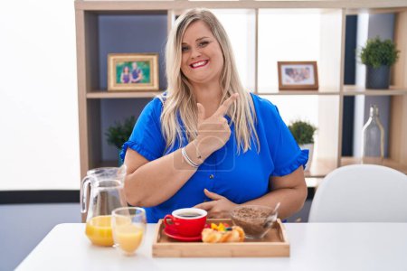 Foto de Caucásico más tamaño mujer desayunando en casa alegre con una sonrisa de la cara señalando con la mano y el dedo hacia arriba a un lado con la expresión feliz y natural en la cara - Imagen libre de derechos