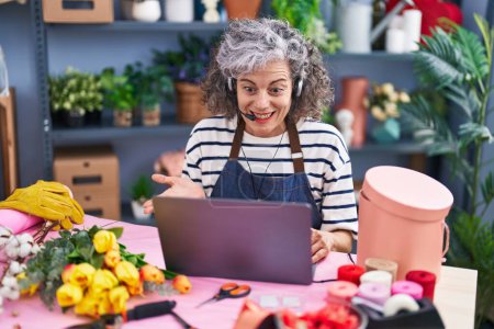 Téléchargez les photos : Femme d'âge moyen avec les cheveux gris travaillant chez le fleuriste avec ordinateur portable célébrant la réalisation avec sourire heureux et expression gagnante avec la main levée - en image libre de droit