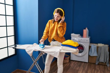Photo for Young blonde woman listening to music ironing clothes at laundry room - Royalty Free Image