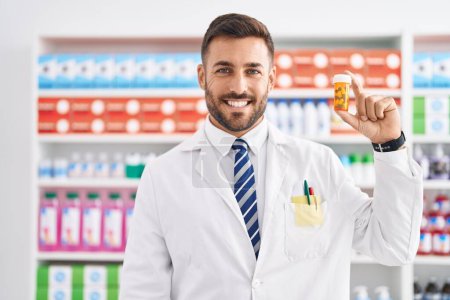 Téléchargez les photos : Bel homme hispanique travaillant à la pharmacie drugstore tenant des pilules regardant positif et heureux debout et souriant avec un sourire confiant montrant des dents - en image libre de droit