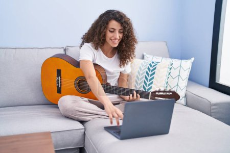 Foto de Young beautiful hispanic woman having online classical guitar class sitting on sofa at home - Imagen libre de derechos