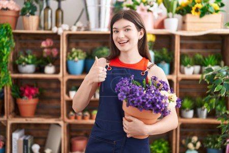 Foto de Mujer caucásica joven que trabaja en floristería sosteniendo la olla con flores sonriendo feliz y positivo, pulgar hacia arriba haciendo excelente y signo de aprobación - Imagen libre de derechos