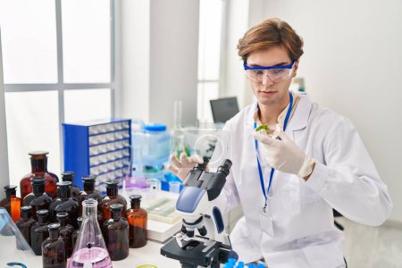 Téléchargez les photos : Young caucasian man scientist using microscope holding plant sample at laboratory - en image libre de droit