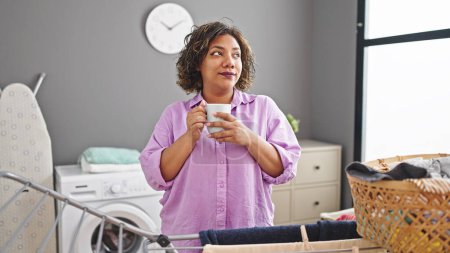 Foto de Joven hermosa mujer latina bebiendo café de pie junto a tendedero en la sala de lavandería - Imagen libre de derechos