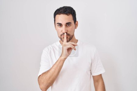 Handsome hispanic man standing over white background asking to be quiet with finger on lips. silence and secret concept. 