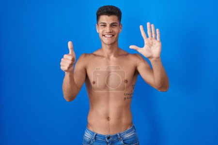 Téléchargez les photos : Jeune homme hispanique debout torse nu sur fond bleu montrant et pointant vers le haut avec les doigts numéro six tout en souriant confiant et heureux. - en image libre de droit