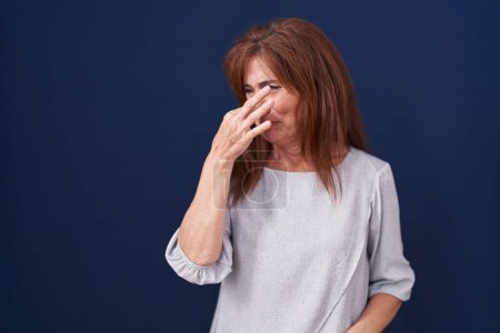 Téléchargez les photos : Femme d'âge moyen debout sur fond bleu sentant quelque chose de puant et dégoûtant, odeur intolérable, retenant souffle avec les doigts sur le nez. mauvaise odeur - en image libre de droit