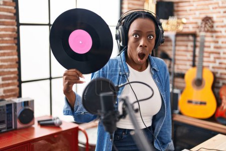 Foto de Hermosa mujer negra sosteniendo disco de vinilo en el estudio de música asustada y sorprendida con la boca abierta para sorpresa, cara de incredulidad - Imagen libre de derechos