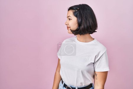 Foto de Mujer hispana joven con camiseta blanca casual sobre fondo rosa mirando hacia un lado, pose de perfil relajado con rostro natural y sonrisa confiada. - Imagen libre de derechos