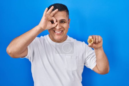 Foto de Joven hispano sosteniendo moneda virtual bitcoin sonriendo feliz haciendo signo ok con la mano en el ojo mirando a través de los dedos - Imagen libre de derechos