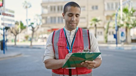 Téléchargez les photos : Jeune homme hispanique volontaire souriant tenant presse-papiers à la rue - en image libre de droit