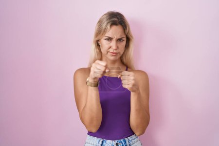 Photo for Young blonde woman standing over pink background ready to fight with fist defense gesture, angry and upset face, afraid of problem - Royalty Free Image