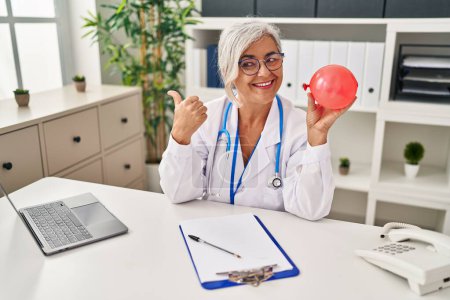 Foto de Mujer de mediana edad con pelo gris vistiendo uniforme médico sosteniendo globo apuntando pulgar hacia el lado sonriendo feliz con la boca abierta - Imagen libre de derechos