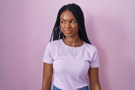 Foto de Mujer afroamericana con trenzas de pie sobre fondo rosa sonriendo mirando hacia un lado y mirando hacia otro pensando. - Imagen libre de derechos